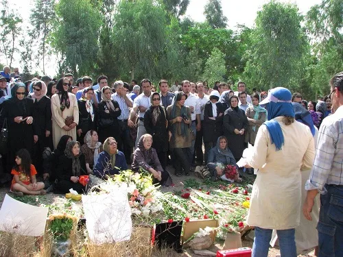 A crowd of mourners standing and sitting around flowers laid on the ground under trees.